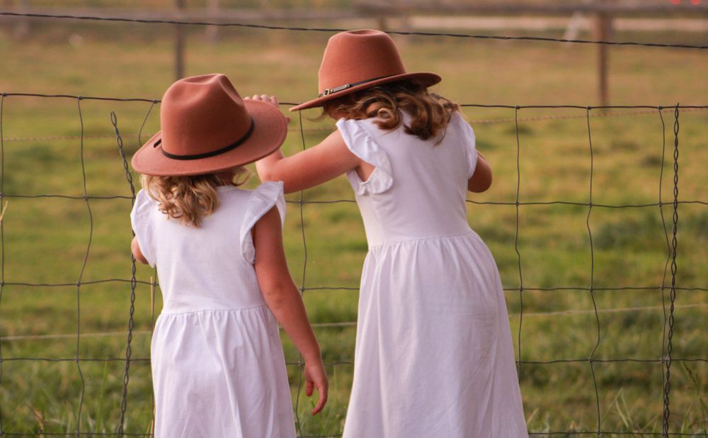 Two children in matching white dresses and brown hats play by a wire fence.