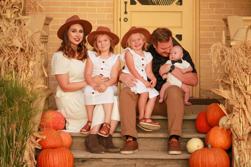 A family sits on brick steps decorated with orange pumpkins for an autumn portrait.