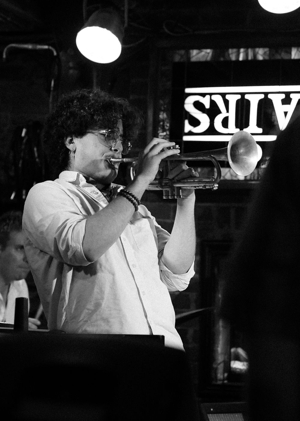 Musician playing trumpet on stage in black and white photo with illuminated sign.