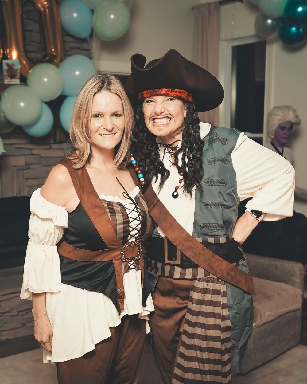 Two people in pirate costumes posing at a party with teal balloons in the background.