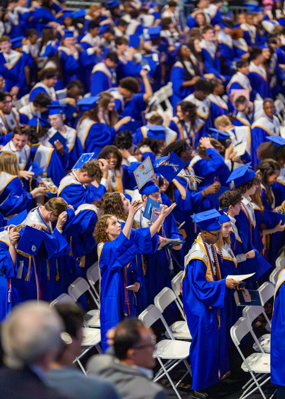 Large crowd of graduates in blue caps and gowns seated at outdoor commencement ceremony.