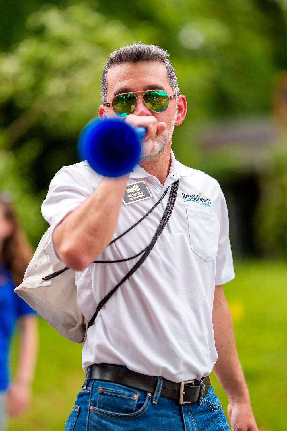 Person in white polo shirt and sunglasses blowing a blue noisemaker in an outdoor setting.