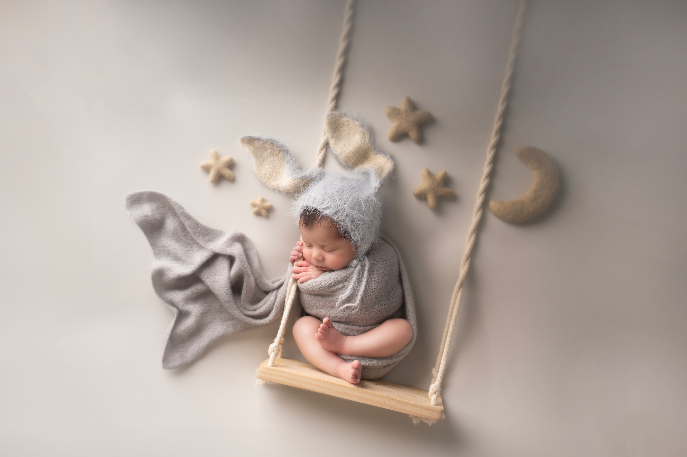 Newborn baby sleeping on a wooden swing with gray bear bonnet, star and moon decorations, soft knit blanket. Embracing Life’s Beauty