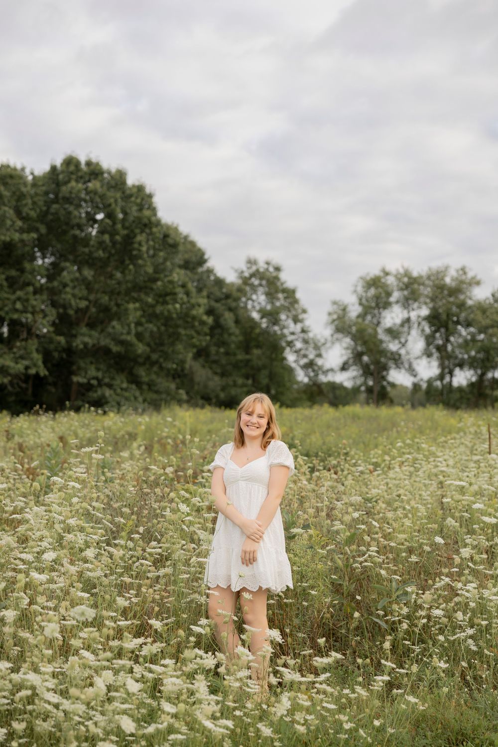 high school senior in a white dress stands joyfully among blooming wildflowers in a summer meadow on an overcast day in lake Orion michigan