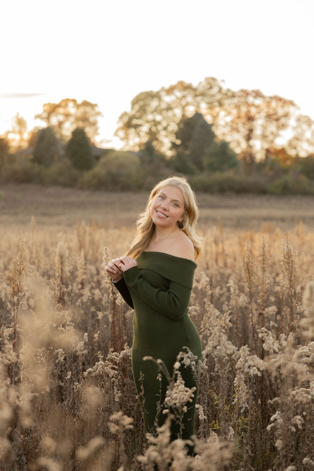 A high school senior in a dark green sweater poses in a golden field of dried wildflowers at sunset in Lake Orion michigan