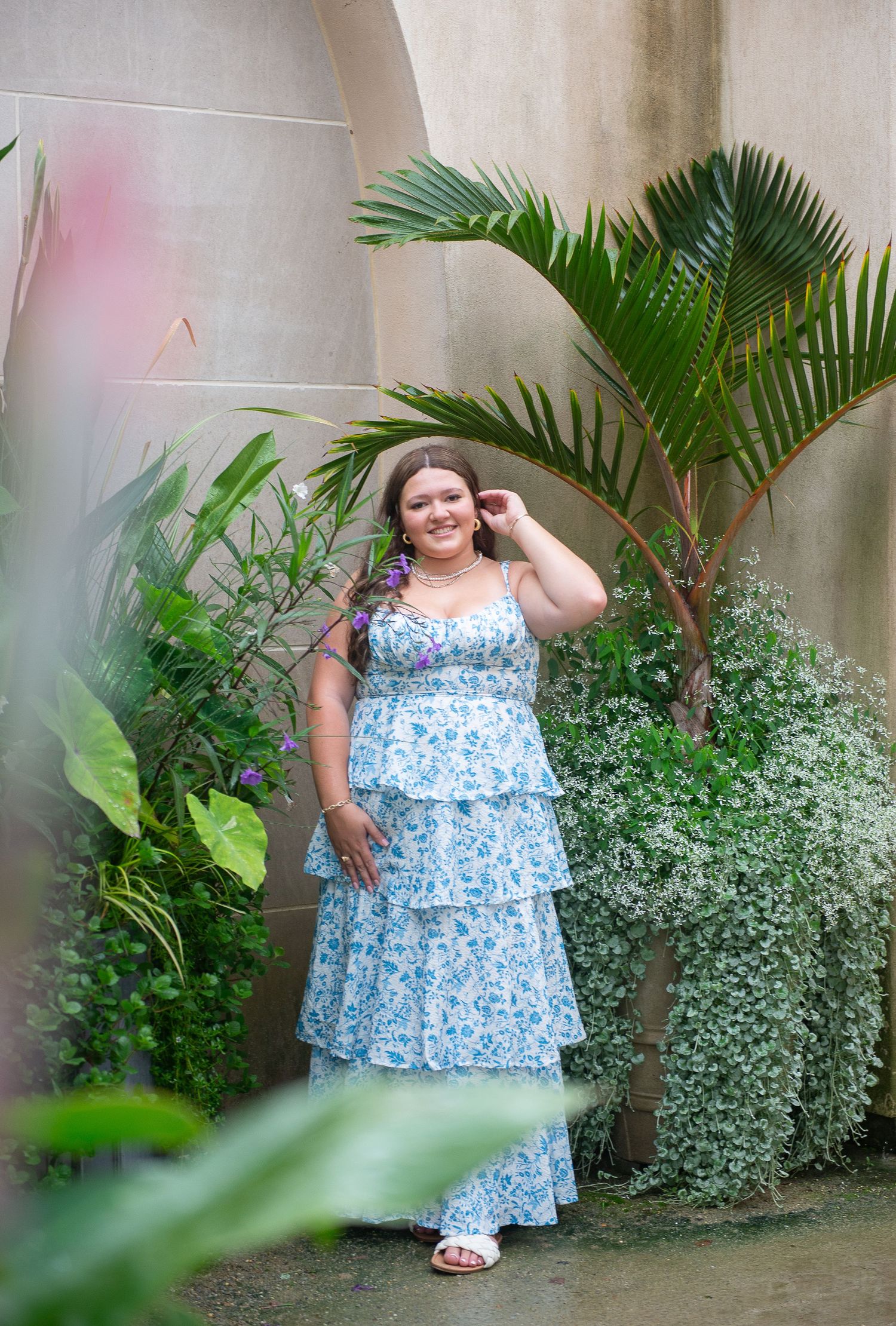 Senior portrait in blue floral dress posing among tropical plants and greenery.