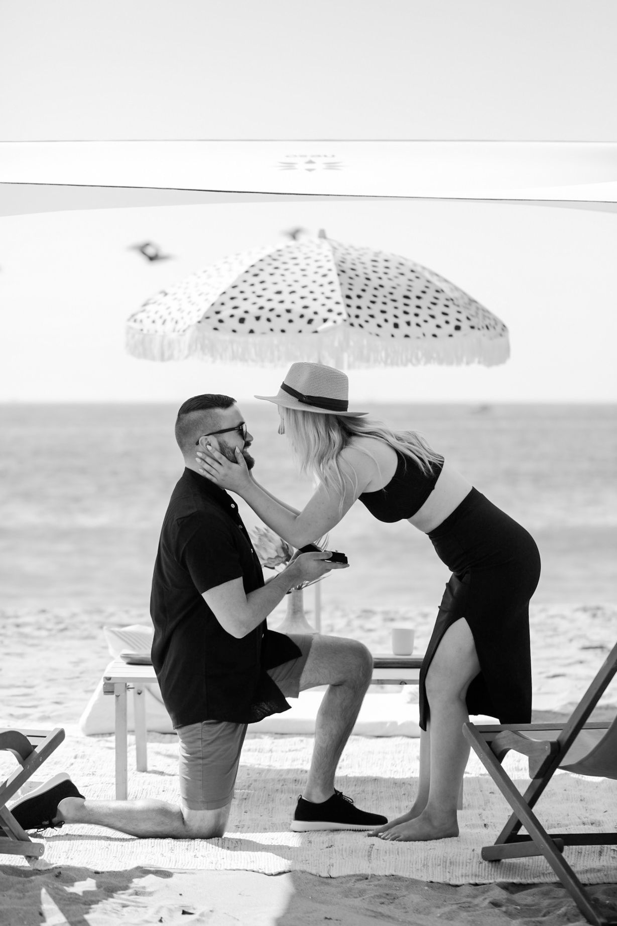 Black and white photo of a beach marriage proposal with man on one knee kissing his partner.