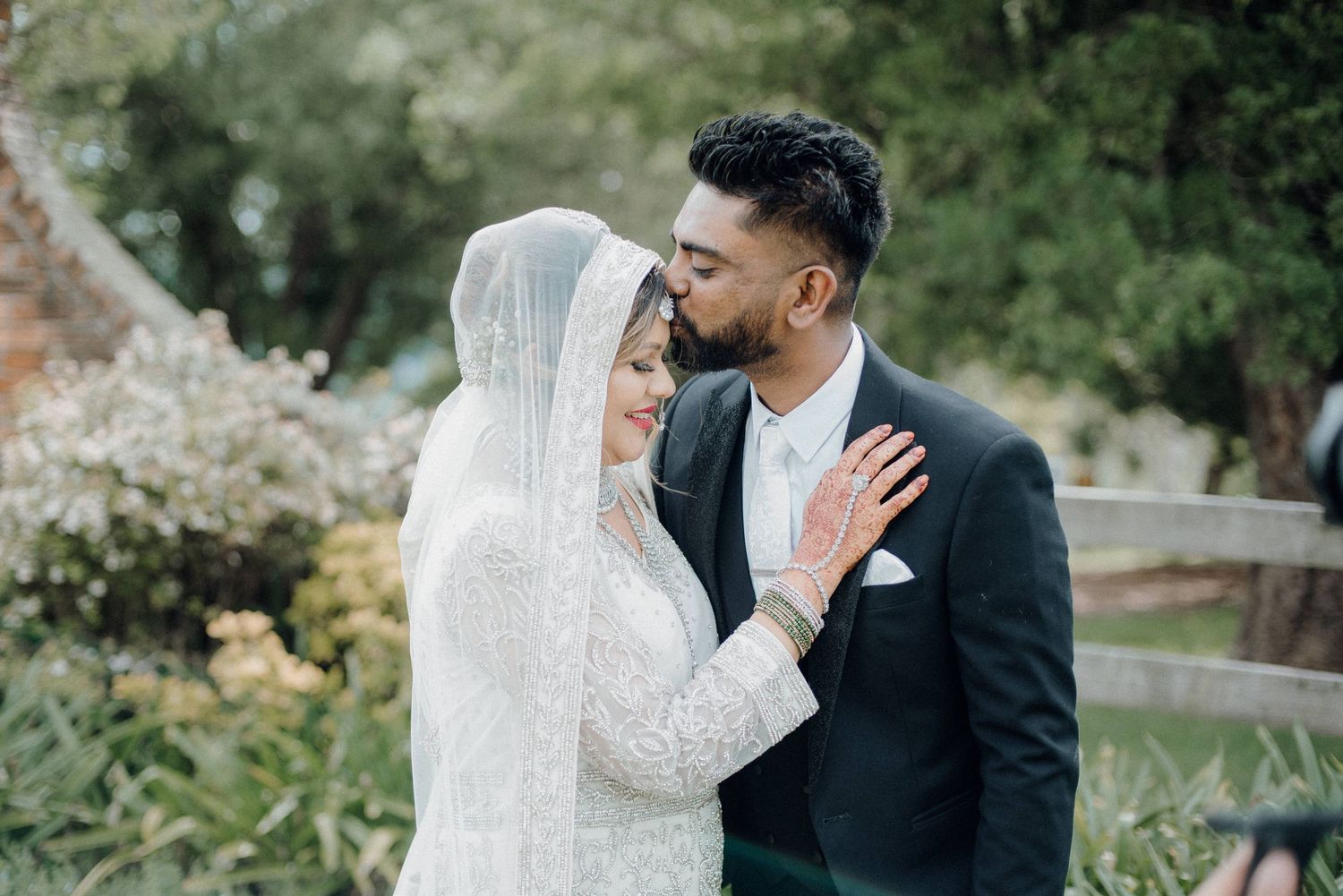 Elegant couple in traditional wedding attire embracing in garden setting.