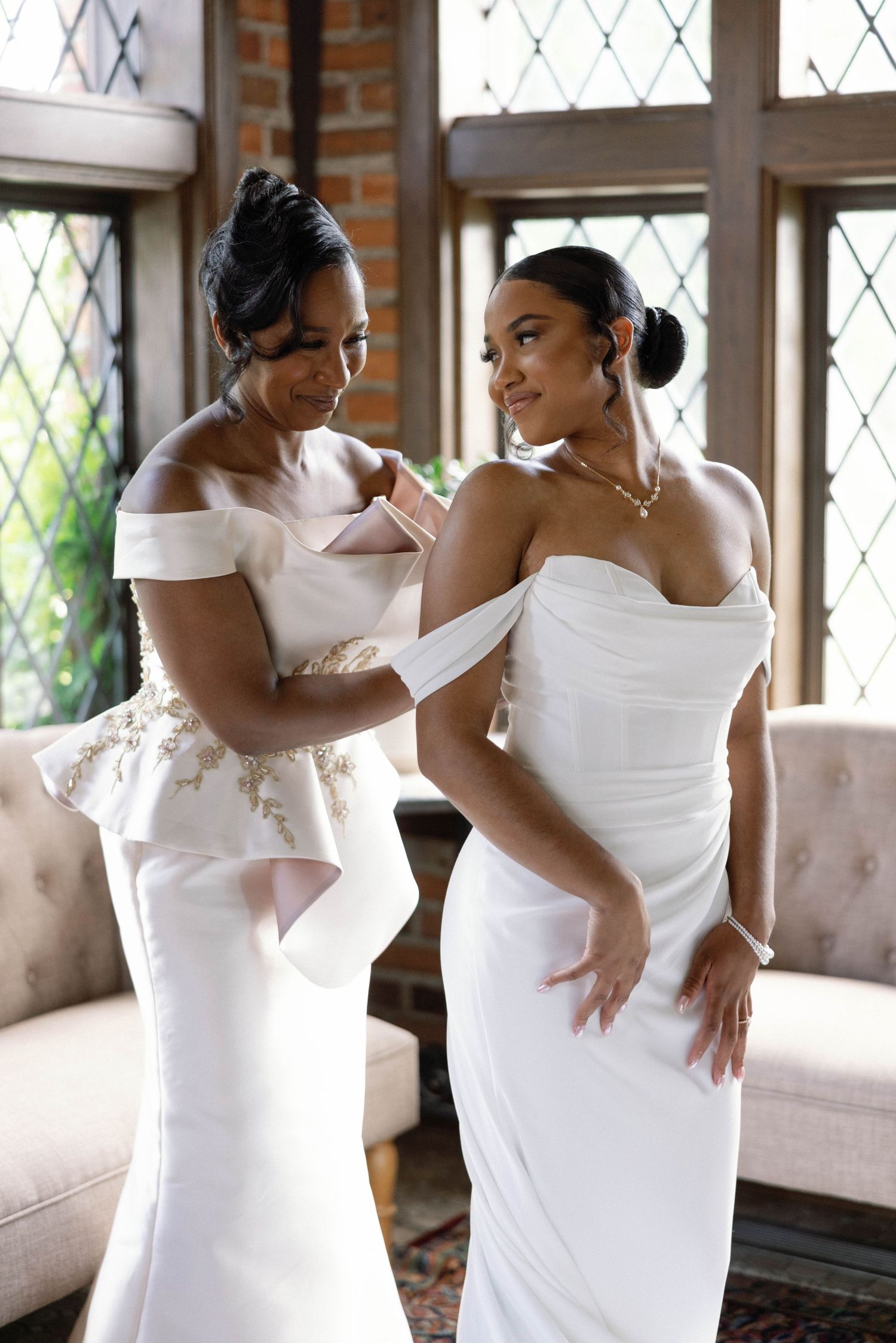 Two women in elegant white dresses help each other prepare for a wedding in a room with vintage windows.