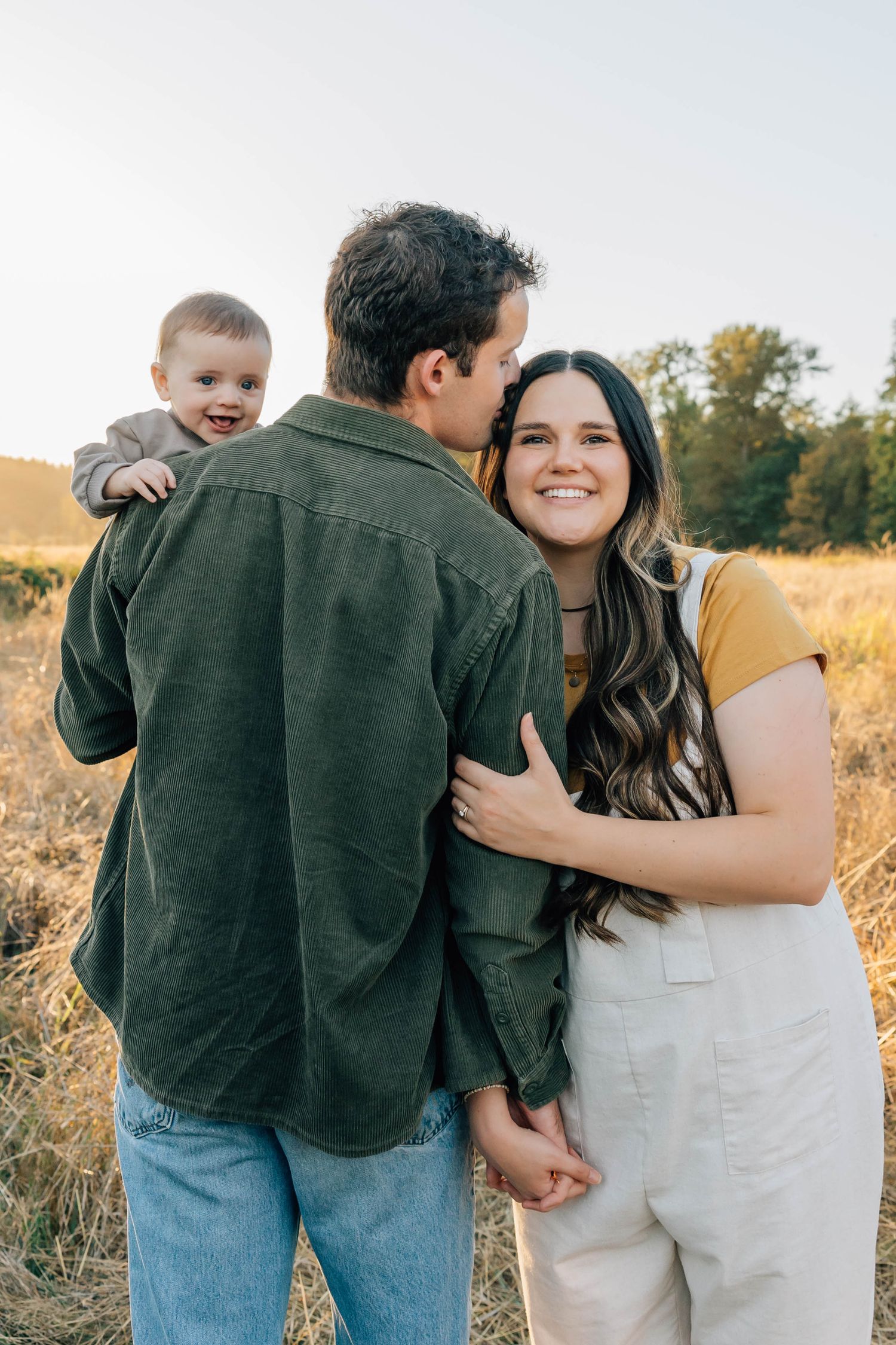 Smiling young mother embraces father holding a happy baby in a golden field.