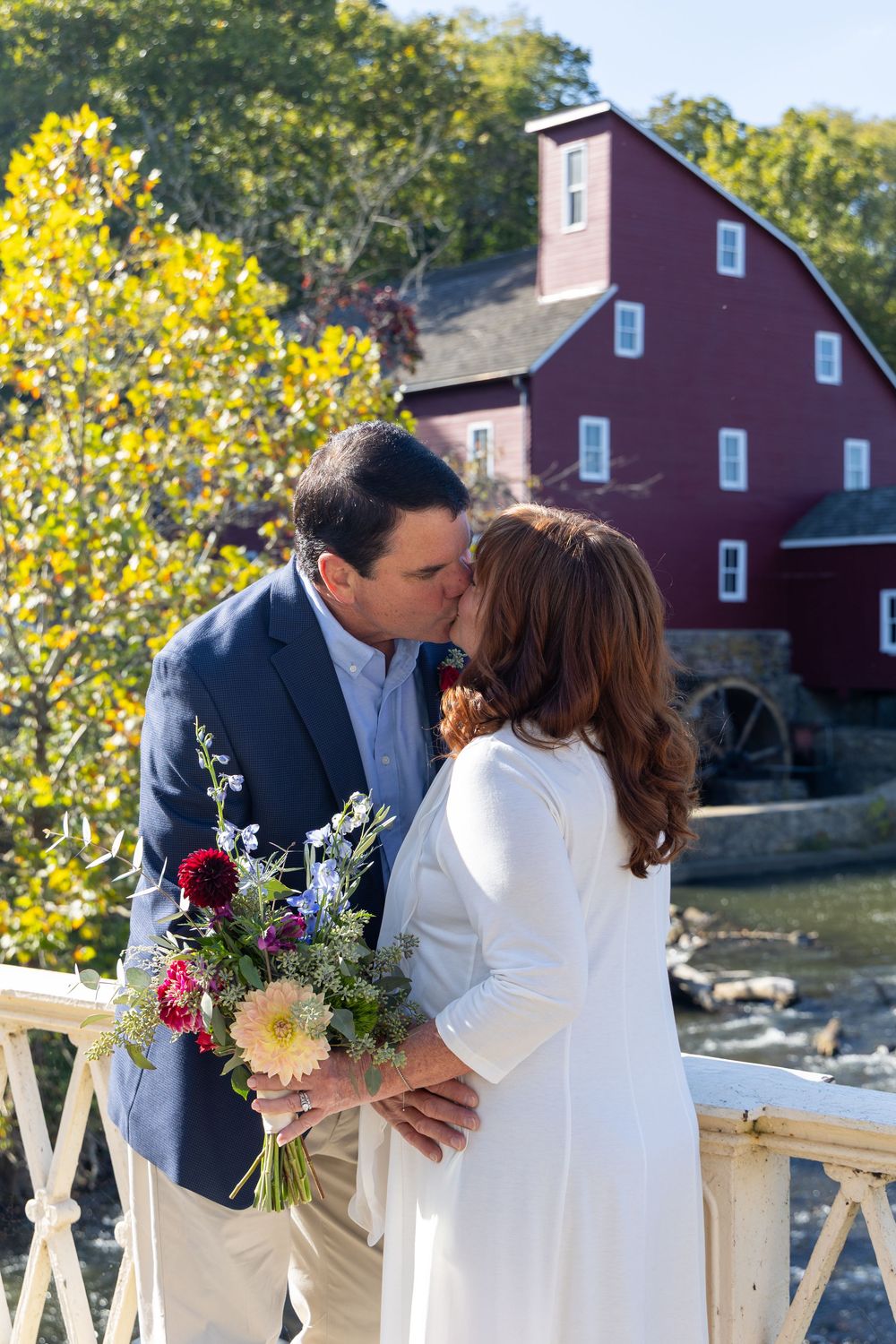 A couple shares intimate moments in front of a rustic red mill building with autumn foliage in the background.