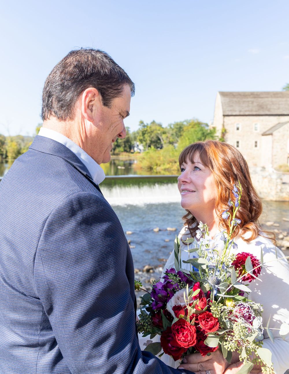 A couple shares a romantic moment by a waterfront stone building while holding a vibrant floral bouquet.