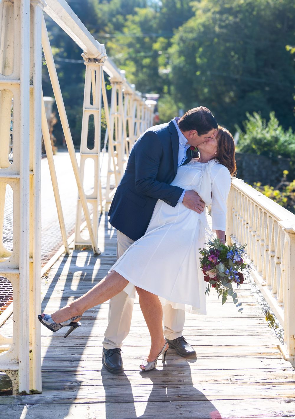 A couple shares a romantic dip kiss on a white wooden bridge in sunlit outdoor setting.