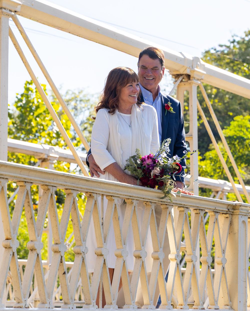 A couple in wedding attire stands on a white balcony railing with decorative bunting during a sunny outdoor celebration.