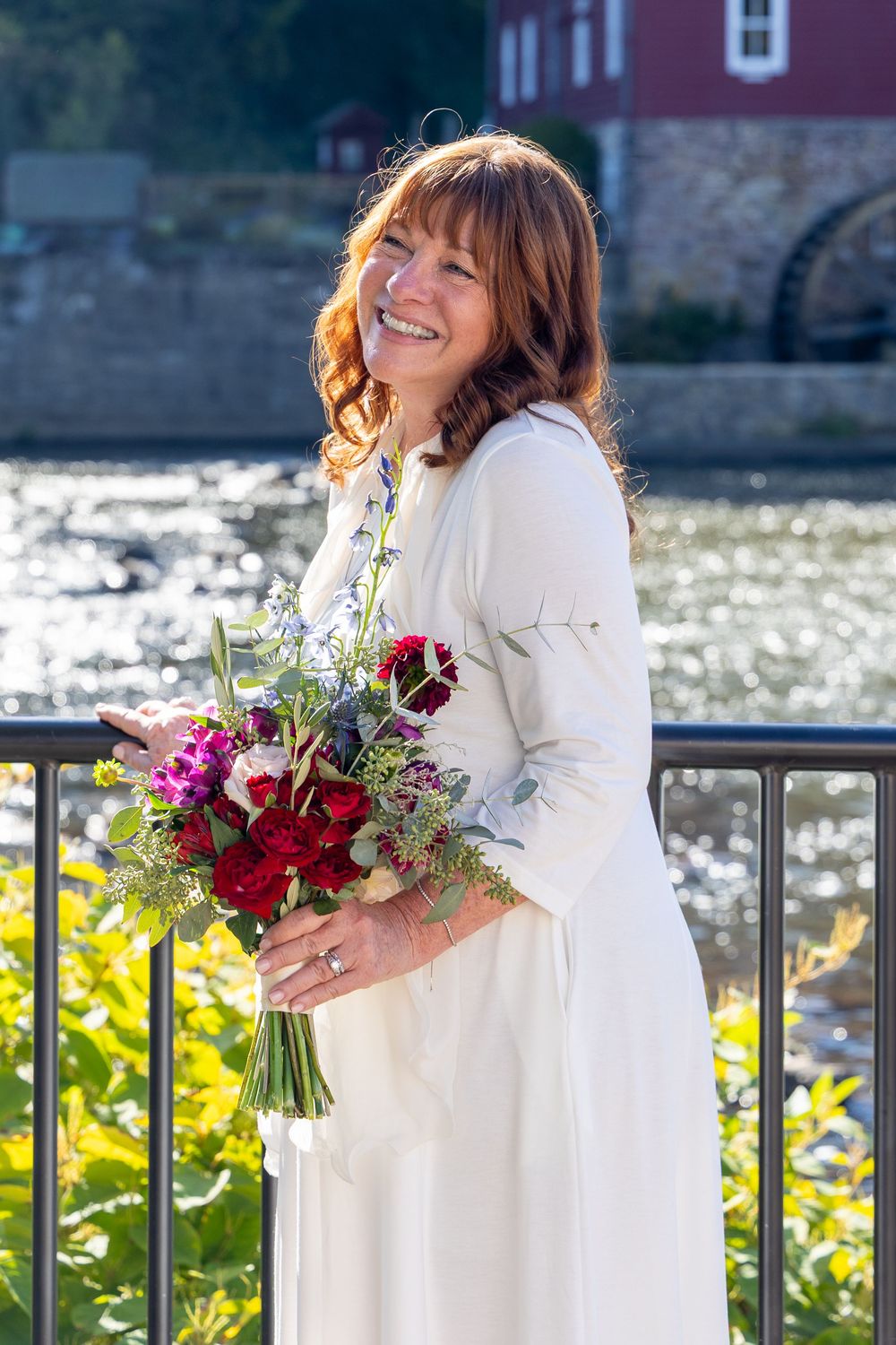 A person in a white dress holds a vibrant bouquet of red and purple flowers against a waterfront railing.