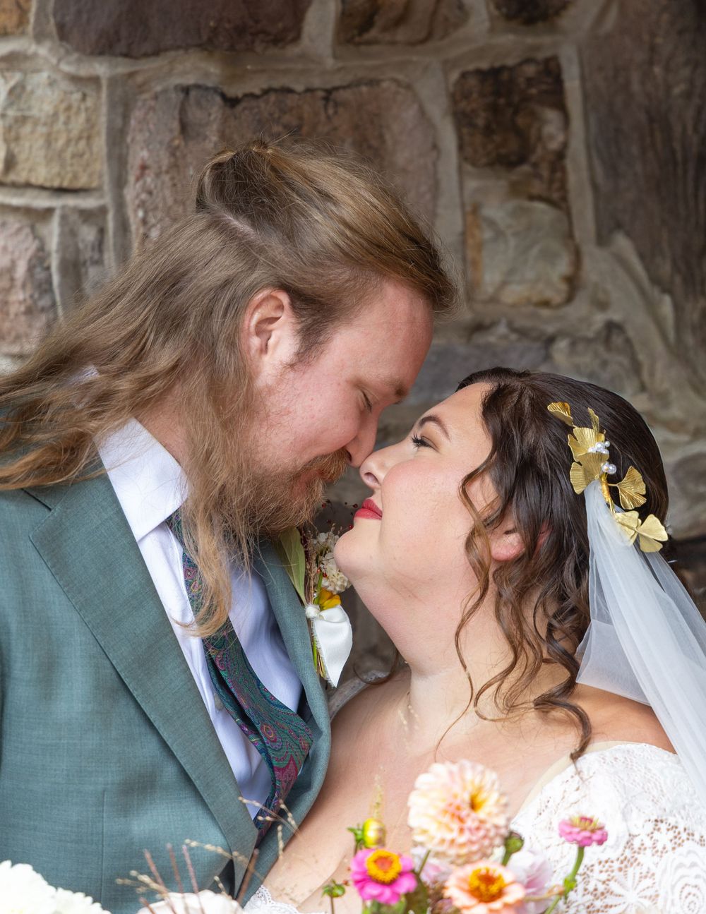 A romantic wedding moment captured against a rustic stone wall with bright flowers in the foreground.