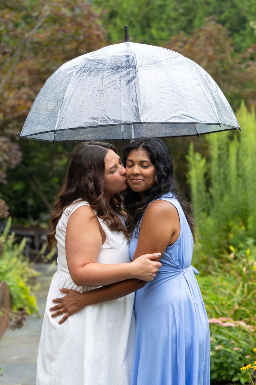 Two women share a tender kiss under a clear umbrella in a lush garden during rain, one in white, one in blue dress.