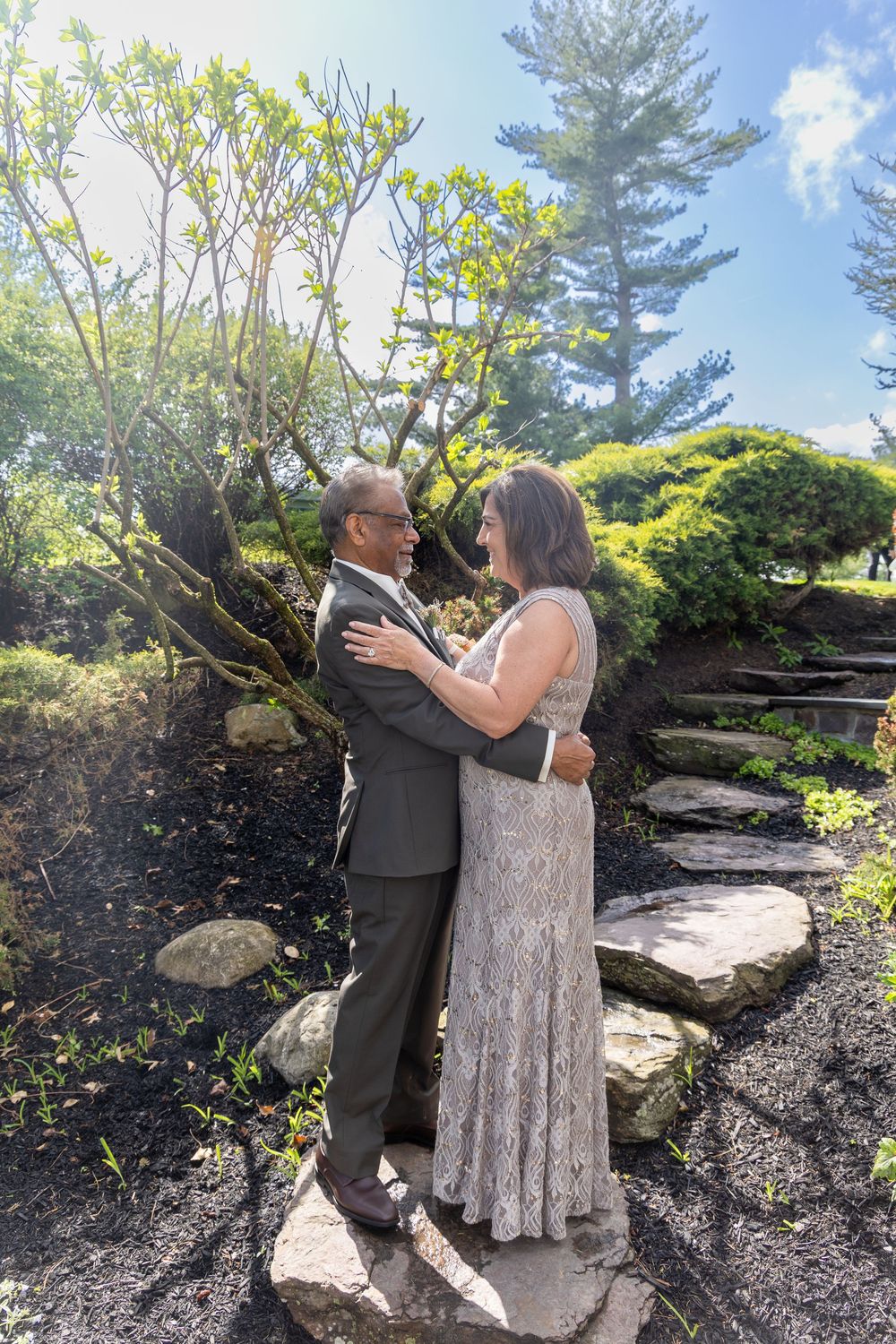 Couple in formal attire embracing in a lush garden with stone steps and trees on a sunny day.