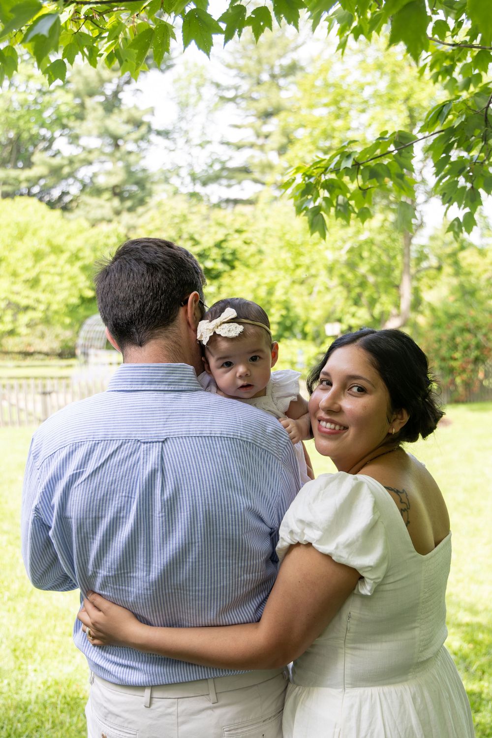 A smiling family with a baby girl in a headband poses outdoors in a lush green park setting.
