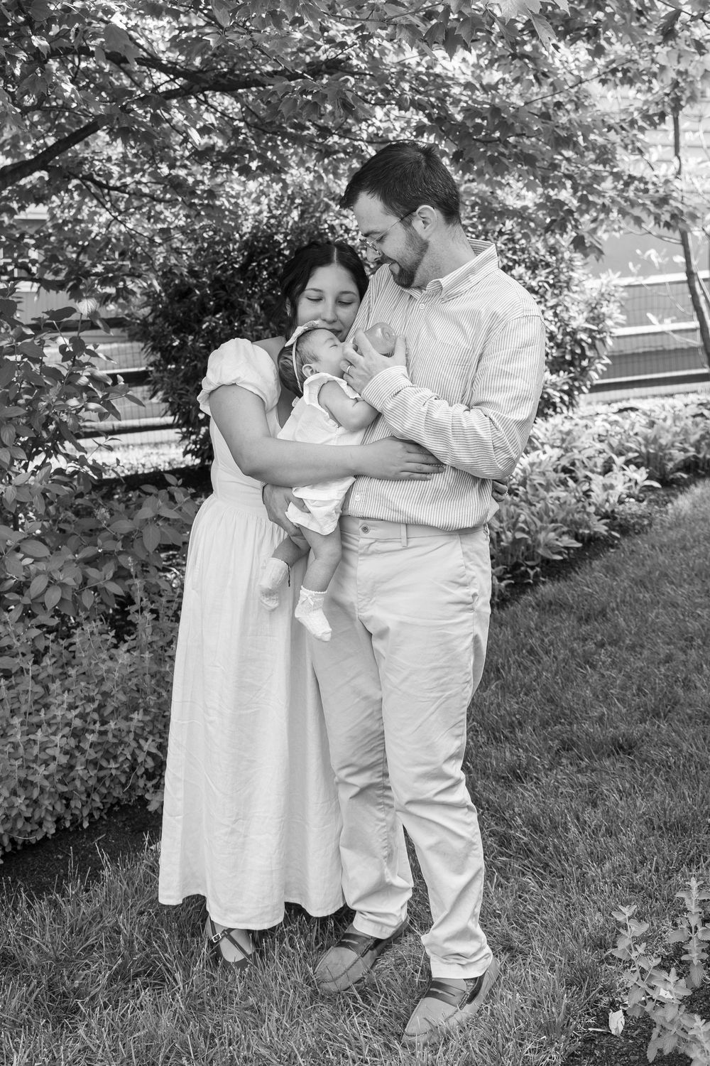 Black and white photo of a couple in white clothing lovingly holding their newborn baby outdoors in a garden.