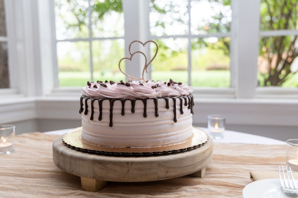 Elegant wedding cake with pink frosting, chocolate drizzle, and heart topper on wooden stand by bright windows.