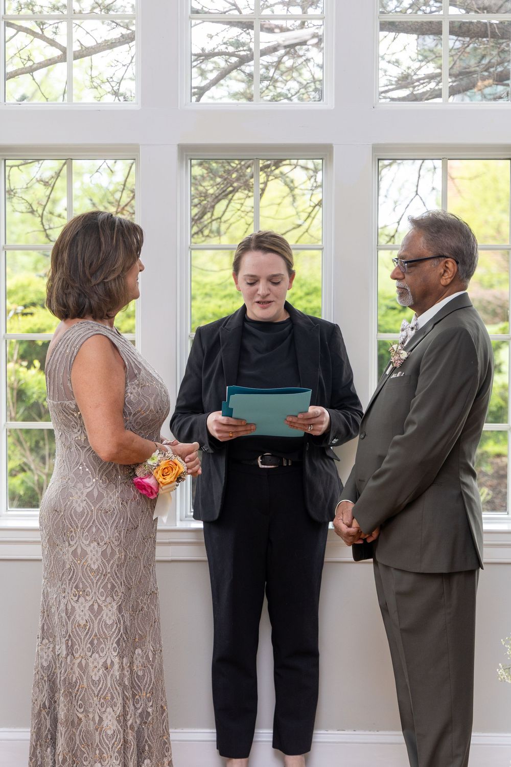 Officiant reads vows to couple during intimate indoor wedding ceremony near large windows with garden view.