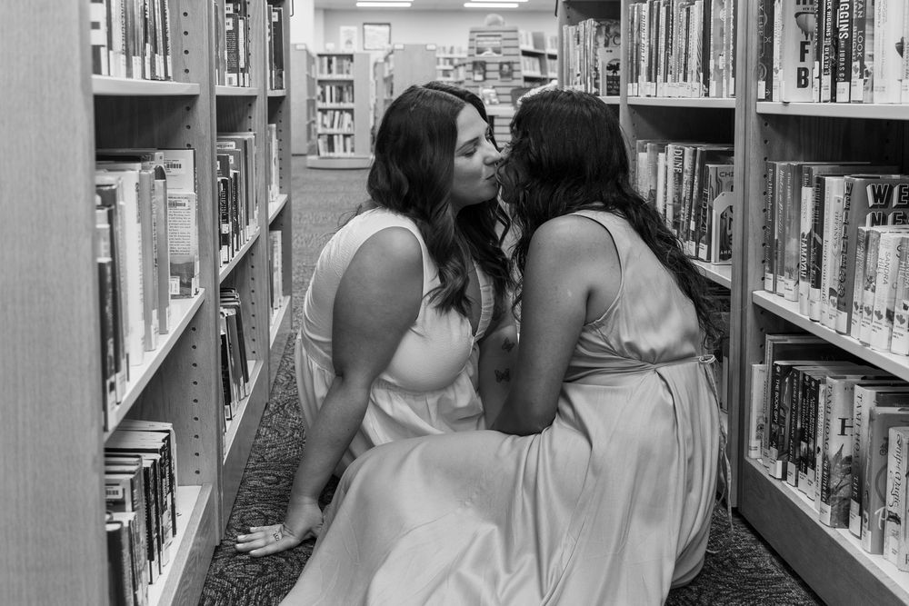 Two brides share a kiss while sitting on the floor between library bookshelves in black and white photo.