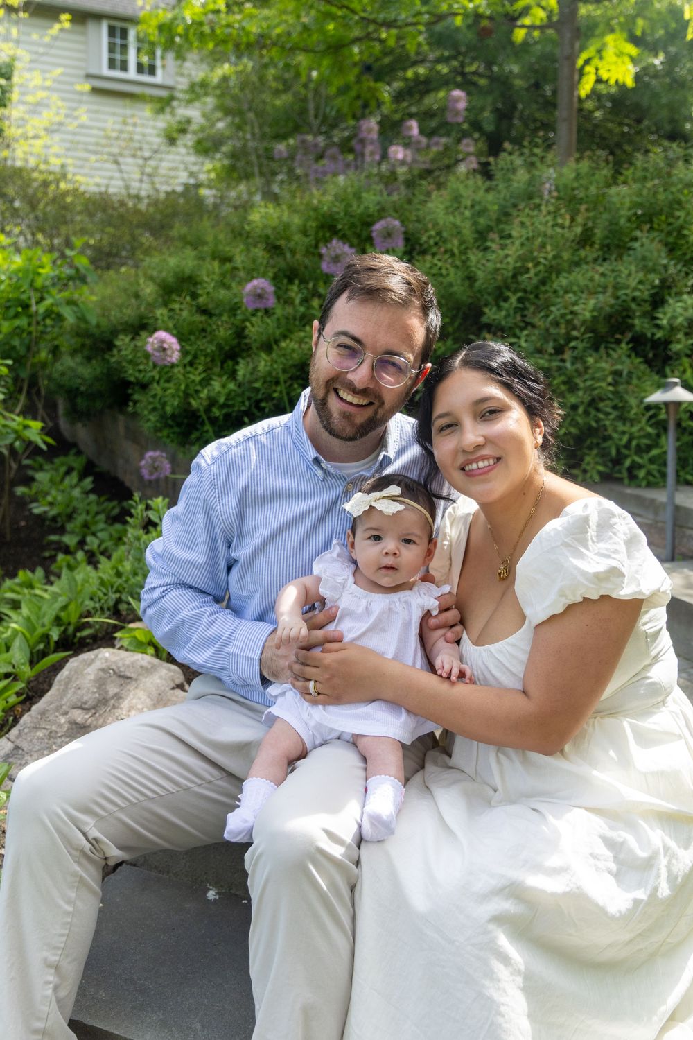Happy family portrait with smiling parents holding their baby girl outdoors in a lush green garden setting.