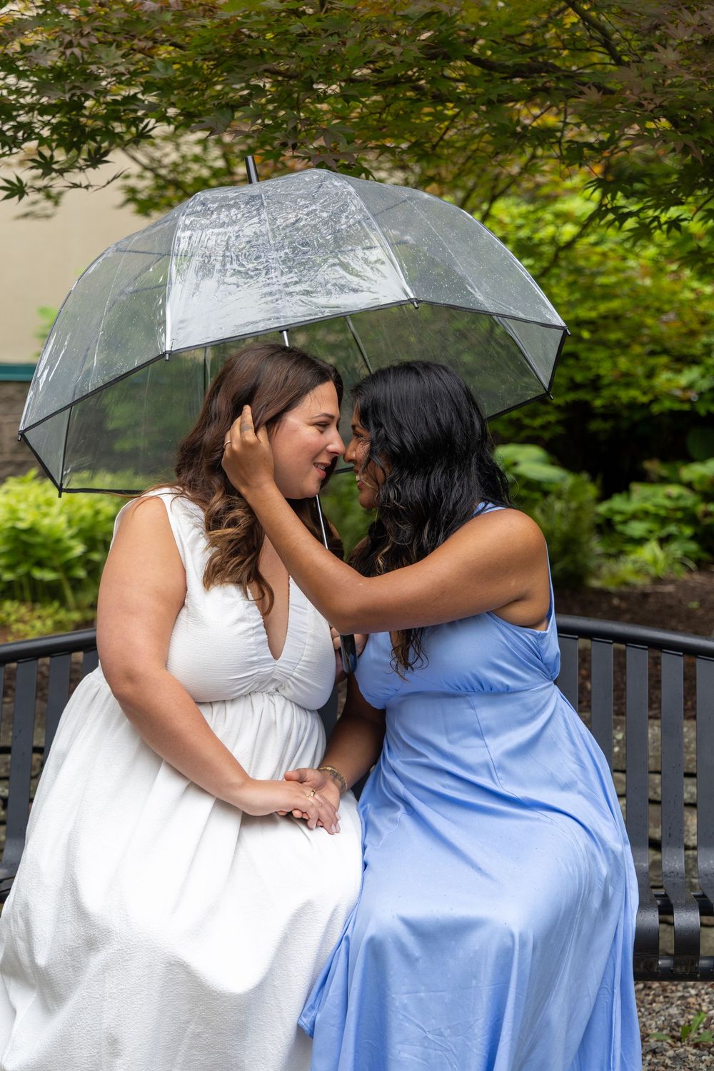 Two brides share a romantic moment under a clear umbrella on a bench, one in white and one in blue, surrounded by lush greenery.