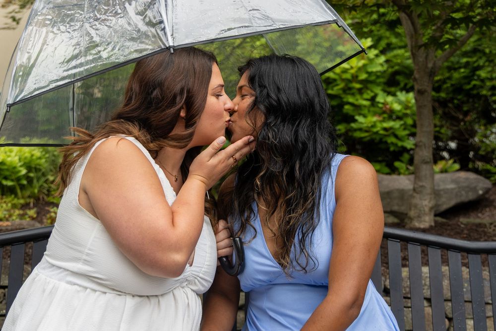Two women sharing a kiss under a clear umbrella in a garden during their wedding.