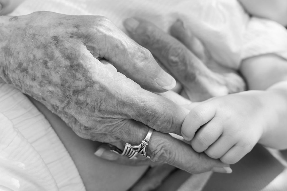 Black and white photo of an elderly person's wrinkled hand with a ring holding a tiny newborn baby's hand.