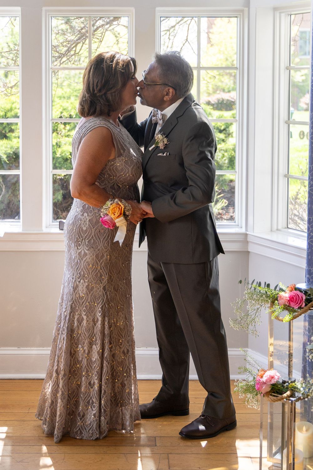 Couple sharing a romantic kiss in elegant wedding attire inside a sunlit room with large windows.