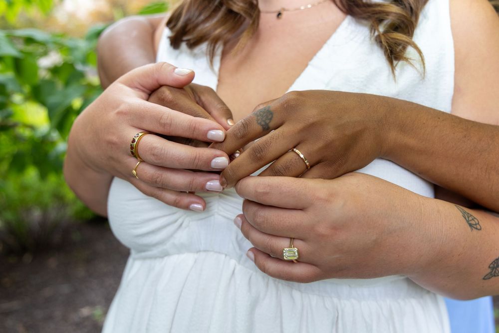 Close-up of two brides holding hands showing engagement rings and wedding bands outdoors.
