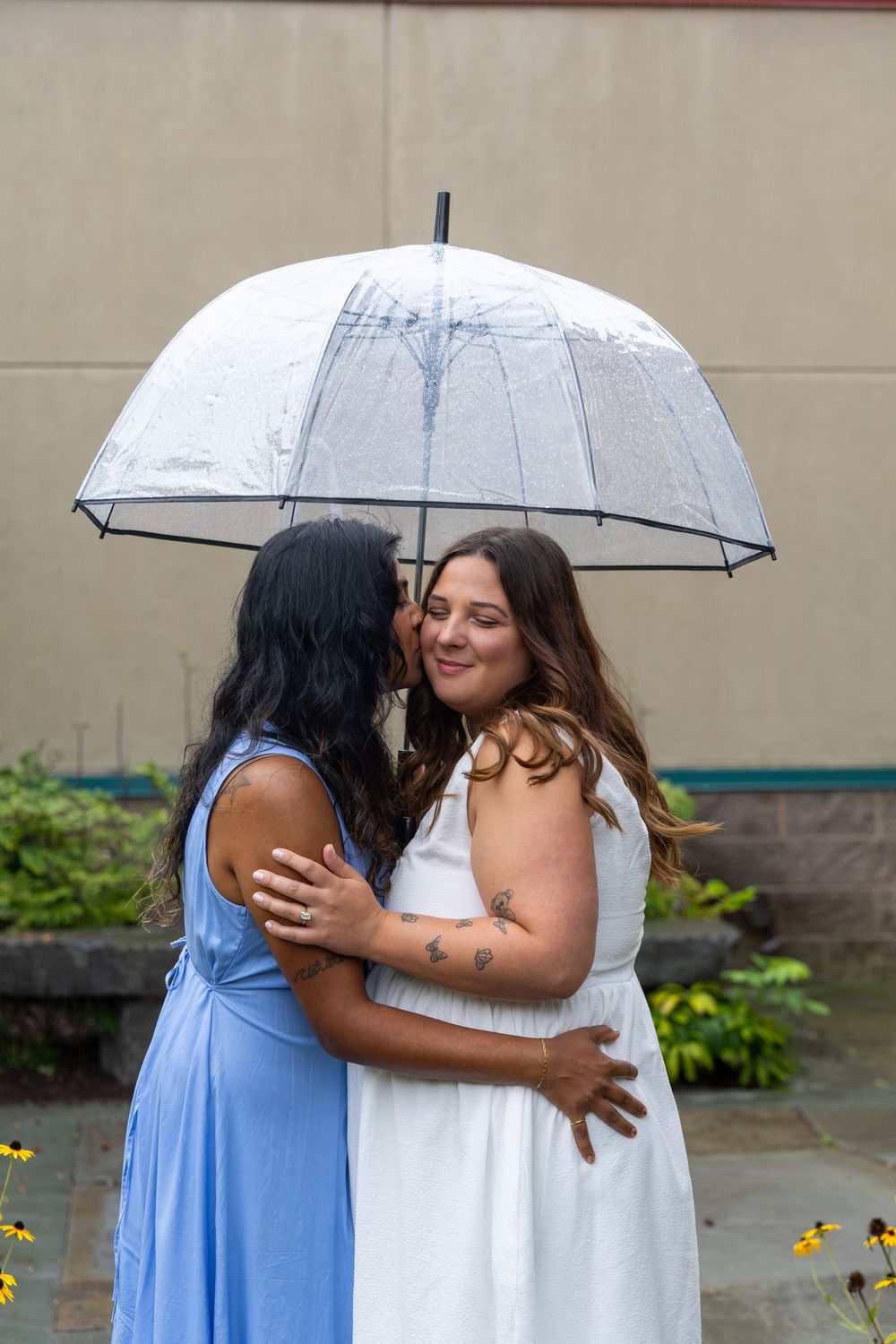 Two women embrace under a clear bubble umbrella in the rain, one in blue dress kissing the other in white.