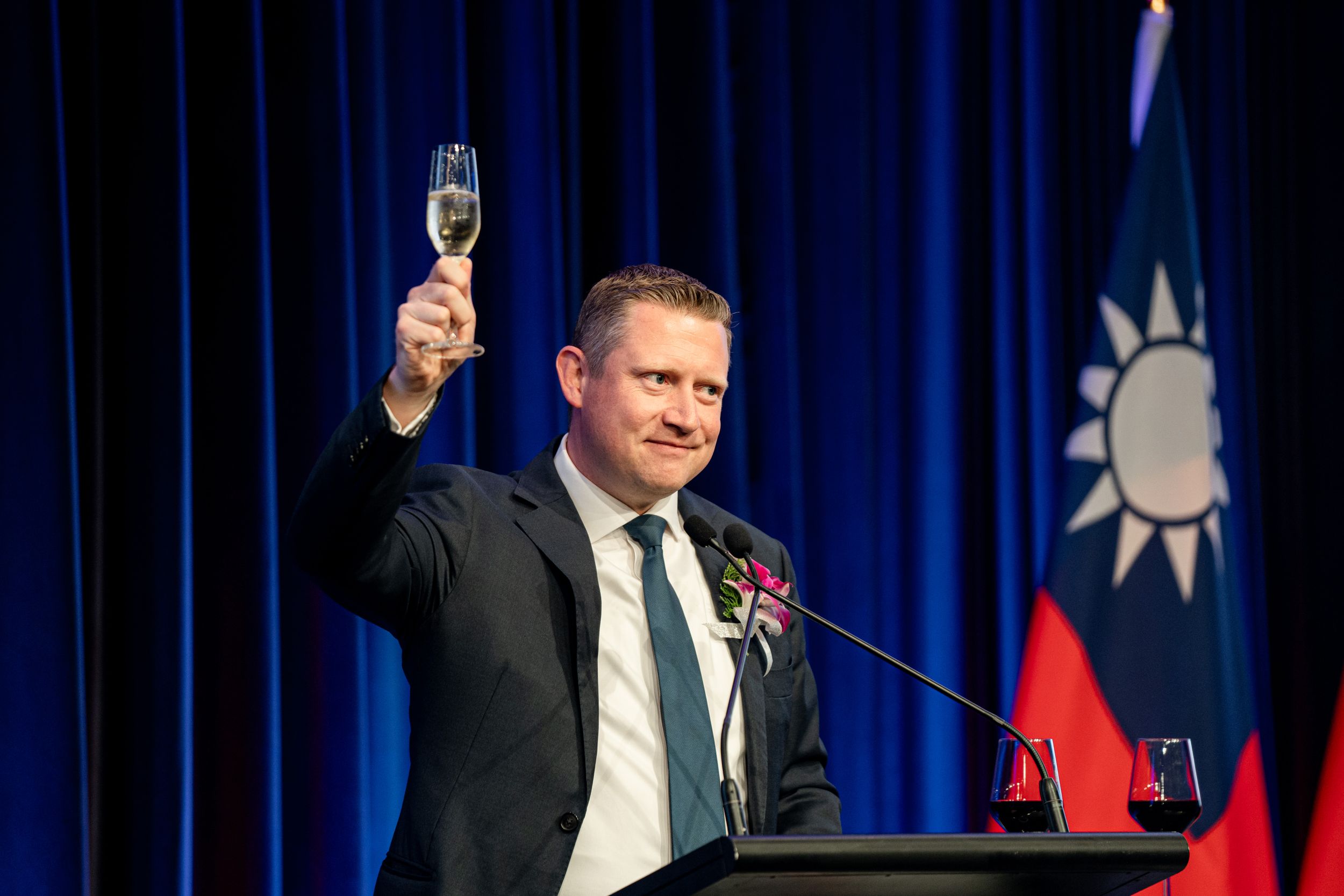 Person in business attire raises a champagne glass during a speech at podium with Taiwan flag backdrop.