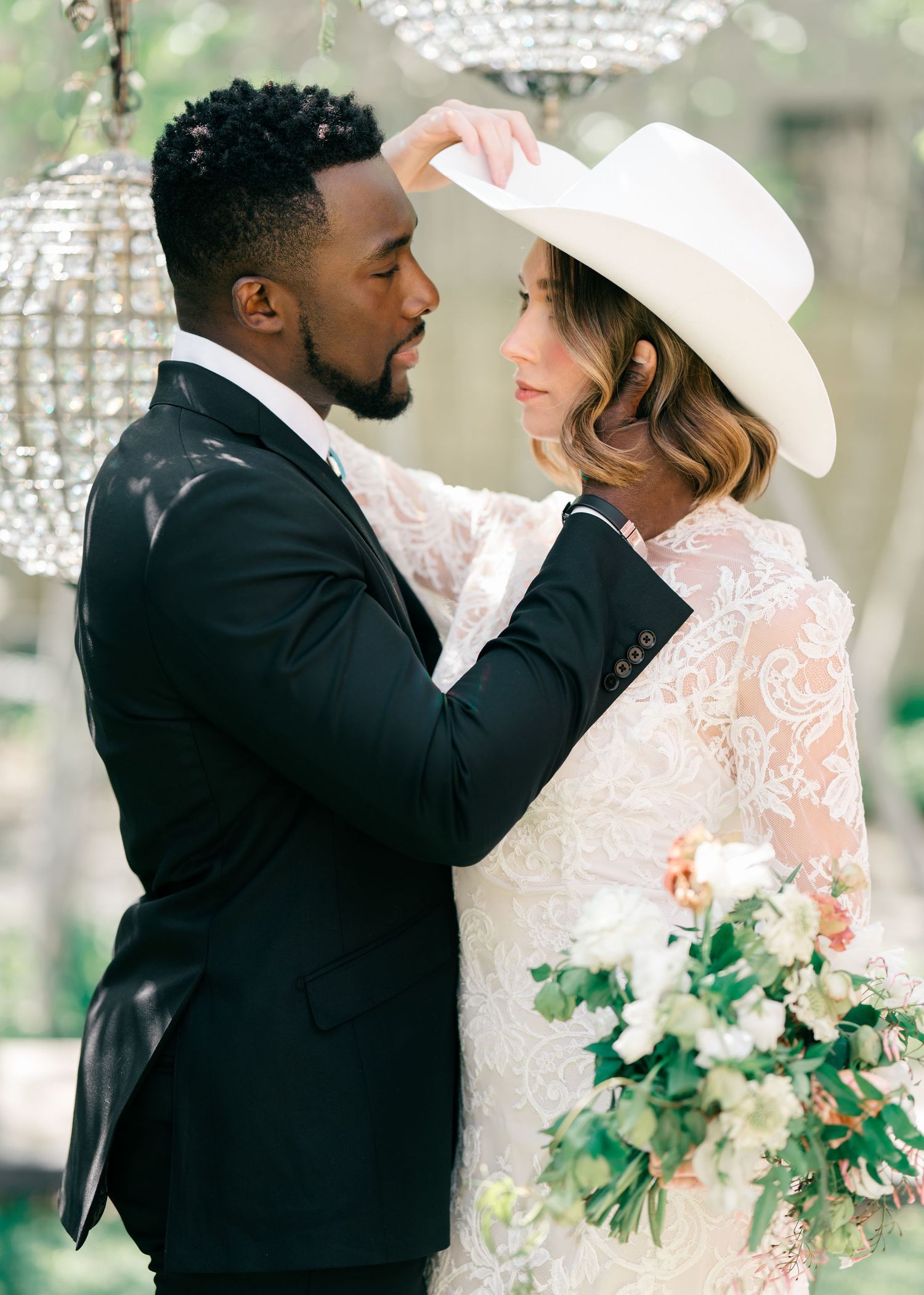 A romantic wedding scene with white lace gown, wide brimmed hat, and black suit against a crystal chandelier backdrop.