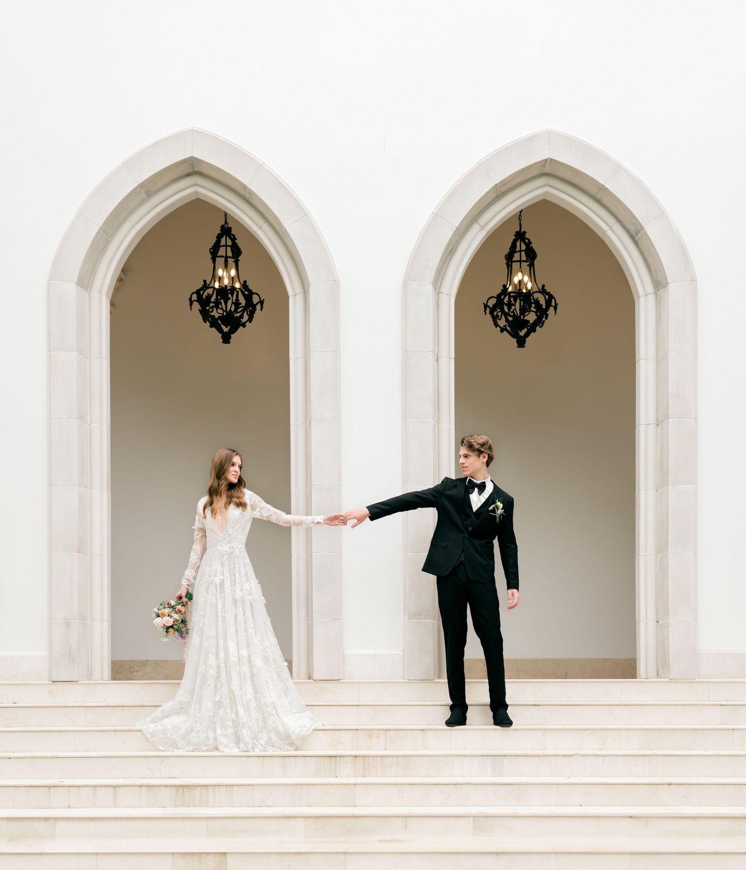 Couple in wedding attire holding hands beneath Gothic arches with hanging chandeliers.