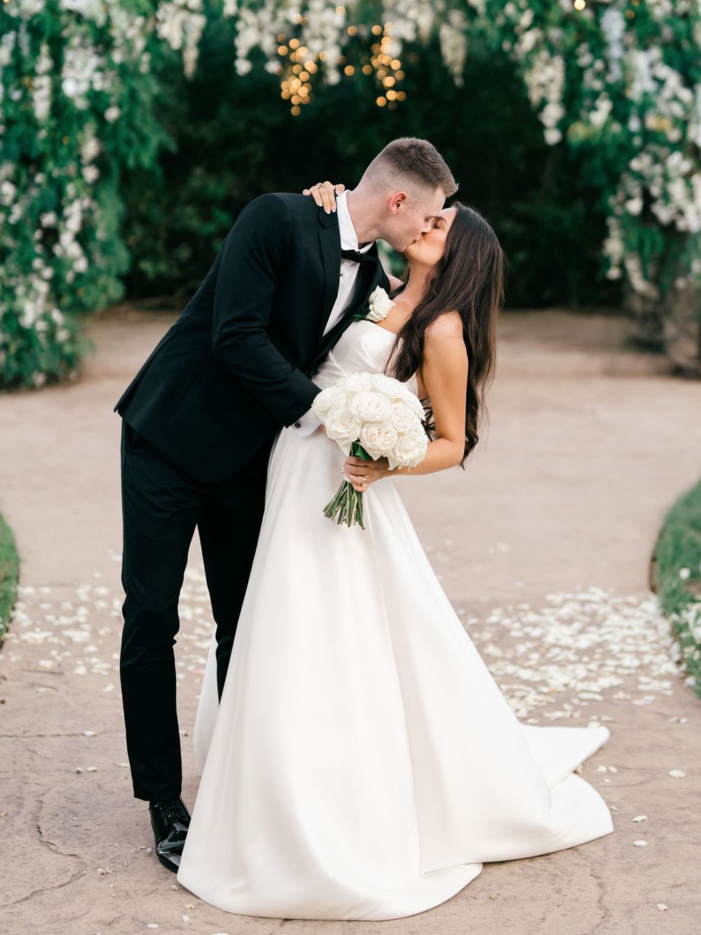 A bride and groom share an intimate dip kiss on their wedding day surrounded by white flower petals on the ground.