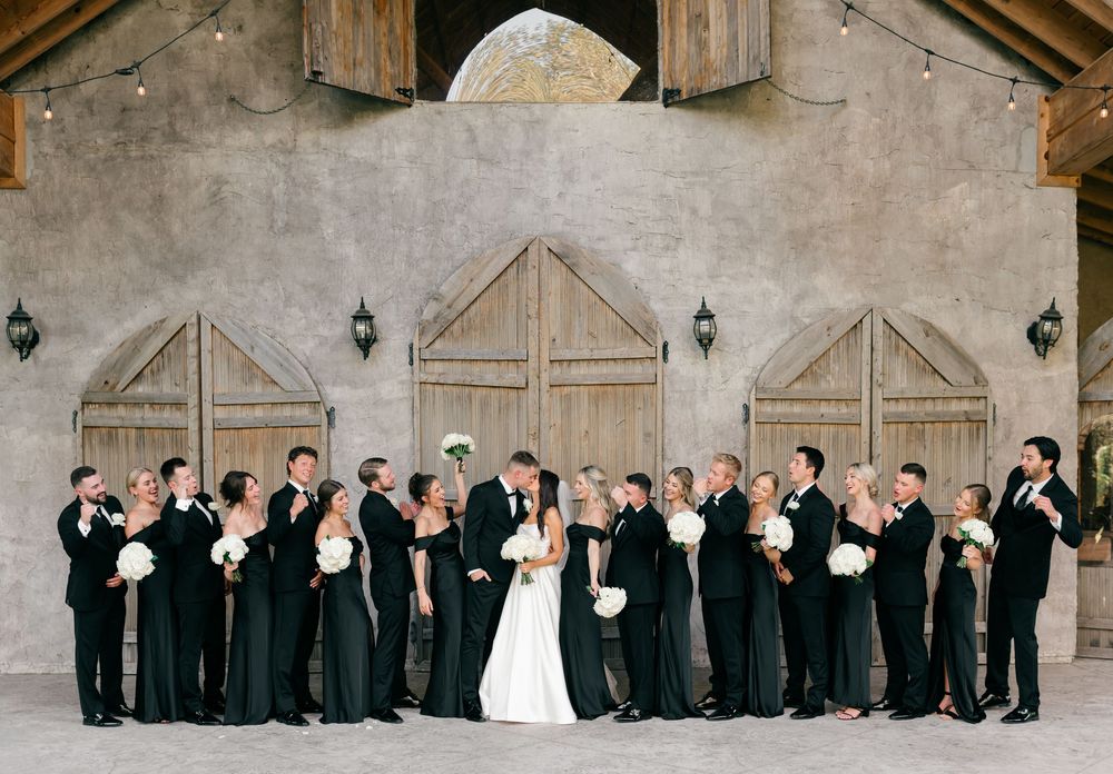 Large wedding party in black attire poses with bride and groom kissing before rustic barn doors.