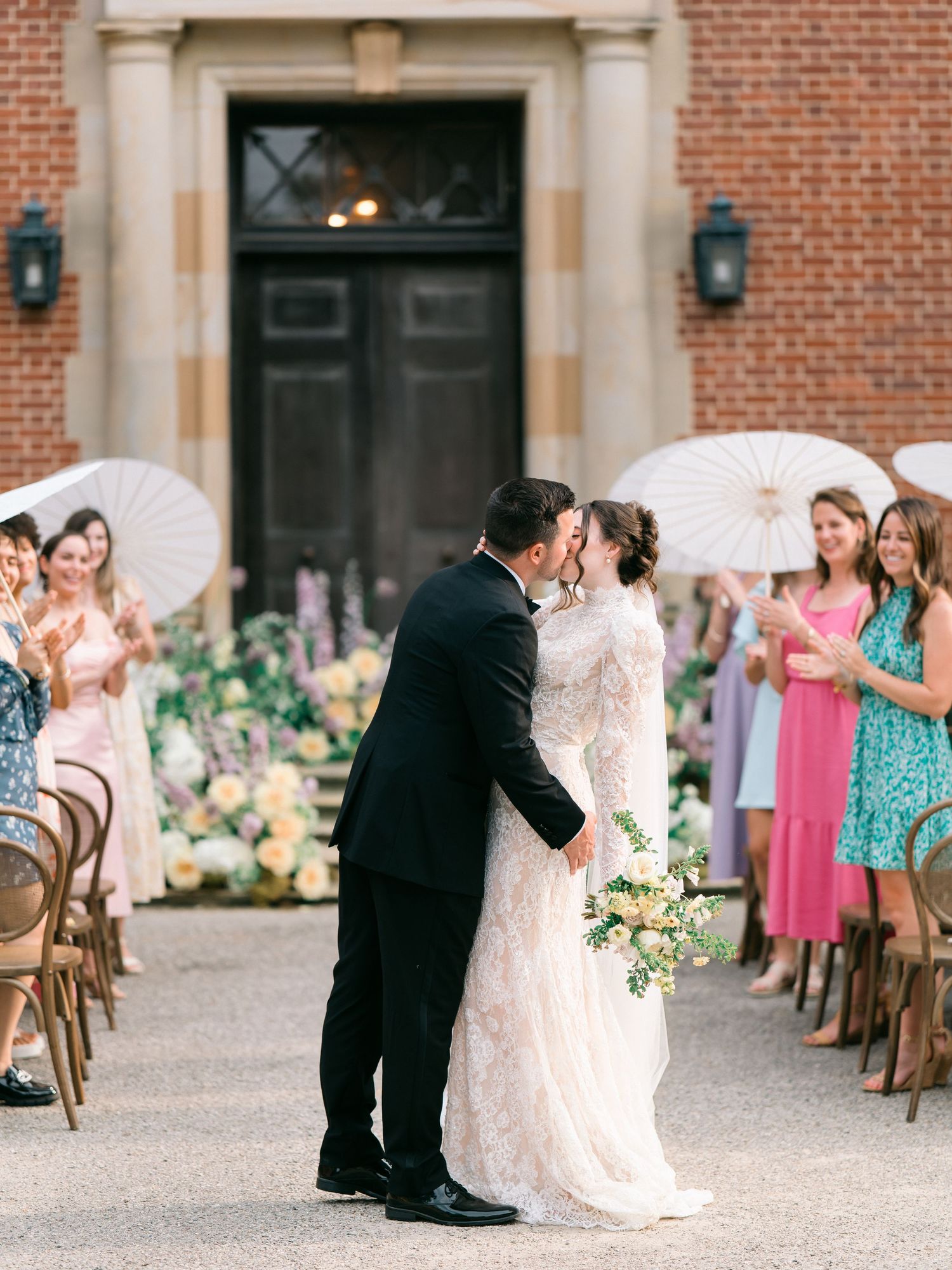 Bride & Groom shares a romantic kiss on brick mansion steps at The Peterloon Estate, while guests with parasols look on at outdoor ceremony.
