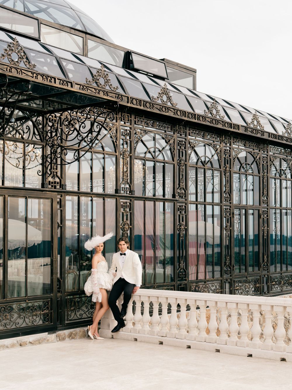 Elegant couple in formal attire posing before ornate Victorian greenhouse with intricate ironwork and glass dome.