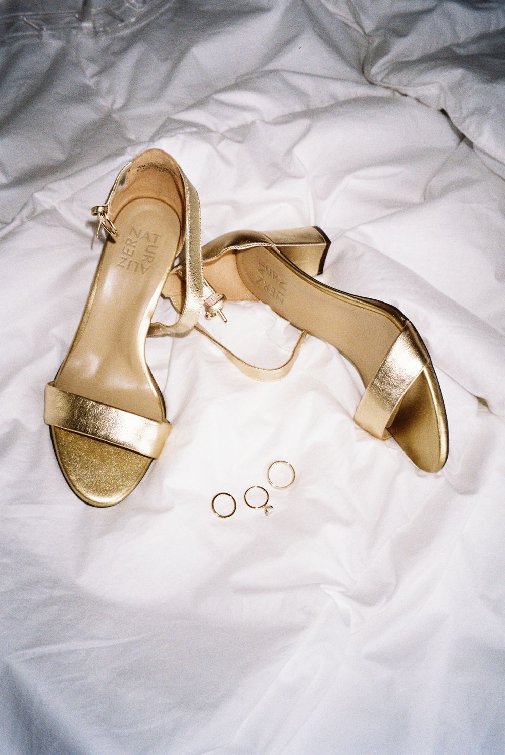 Gold wedding sandals and rings arranged on white fabric.