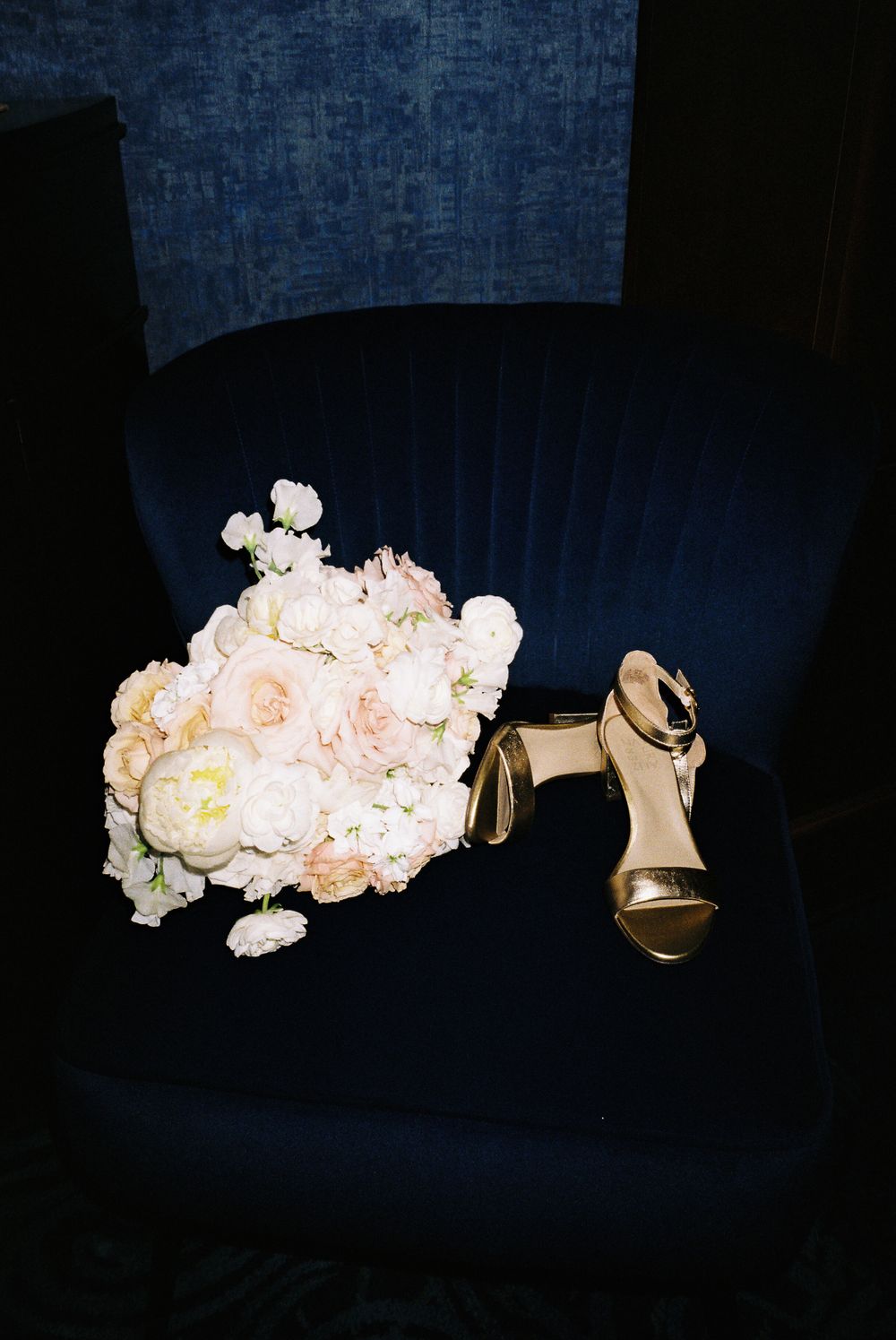 Bridal bouquet of pale pink roses and a gold heel placed on deep blue velvet chair.