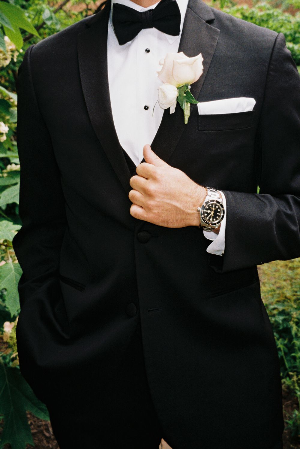 Close-up of groom's classic black tuxedo with white boutonniere and luxury watch.