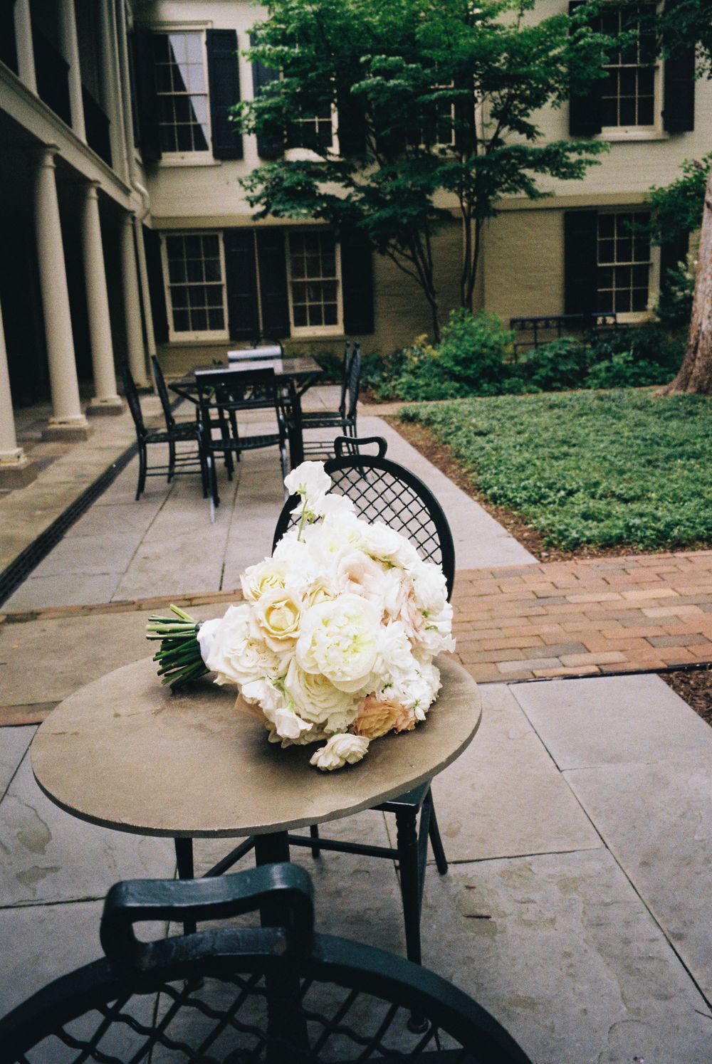 Large white peony bouquet resting on wooden chair in courtyard with brick pavers.