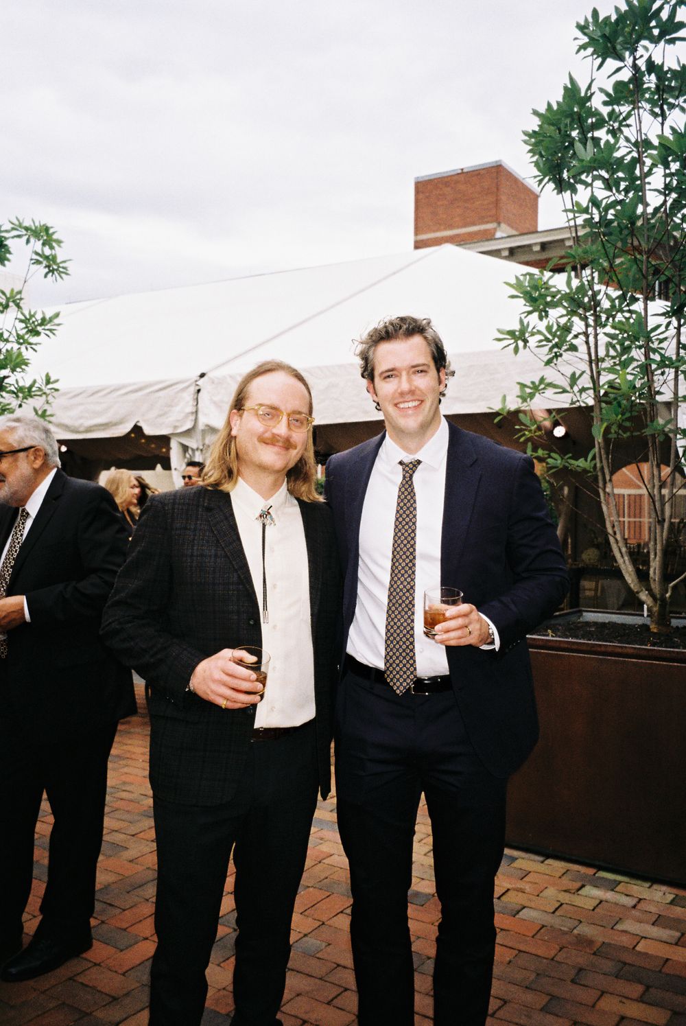 Two wedding guests in formal attire share drinks under a white tent on a brick patio.