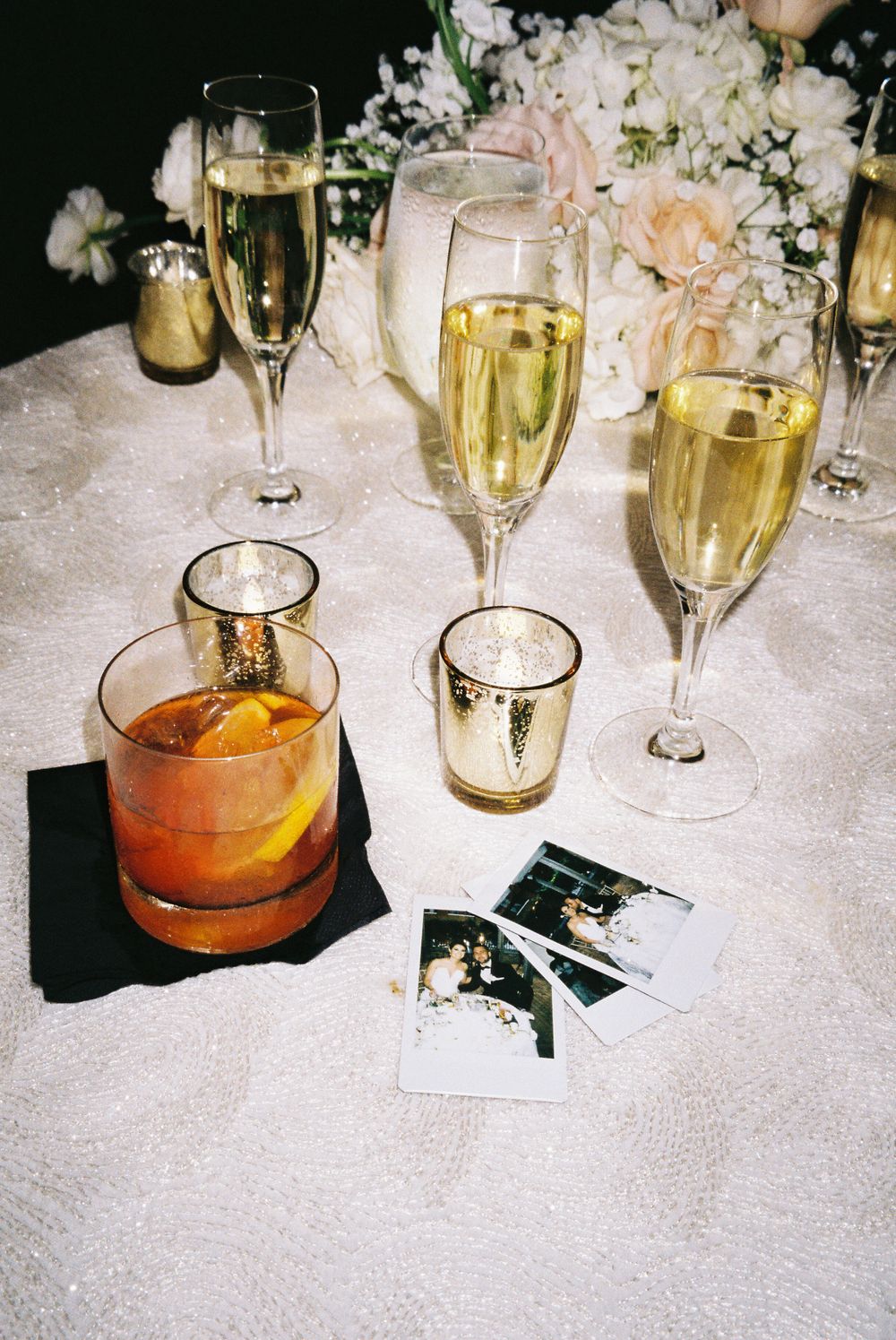 Wedding reception table with champagne flutes, cocktails, and polaroid photos on white tablecloth.