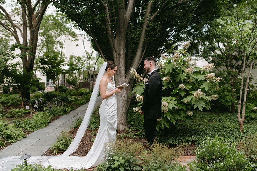 Wedding couple stands beside tree in lush garden setting with long flowing veil detail.