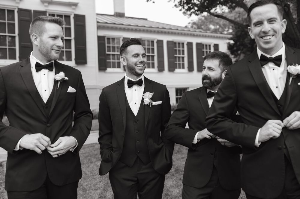 Black and white photo of groomsmen in tuxedos laughing together outdoors.