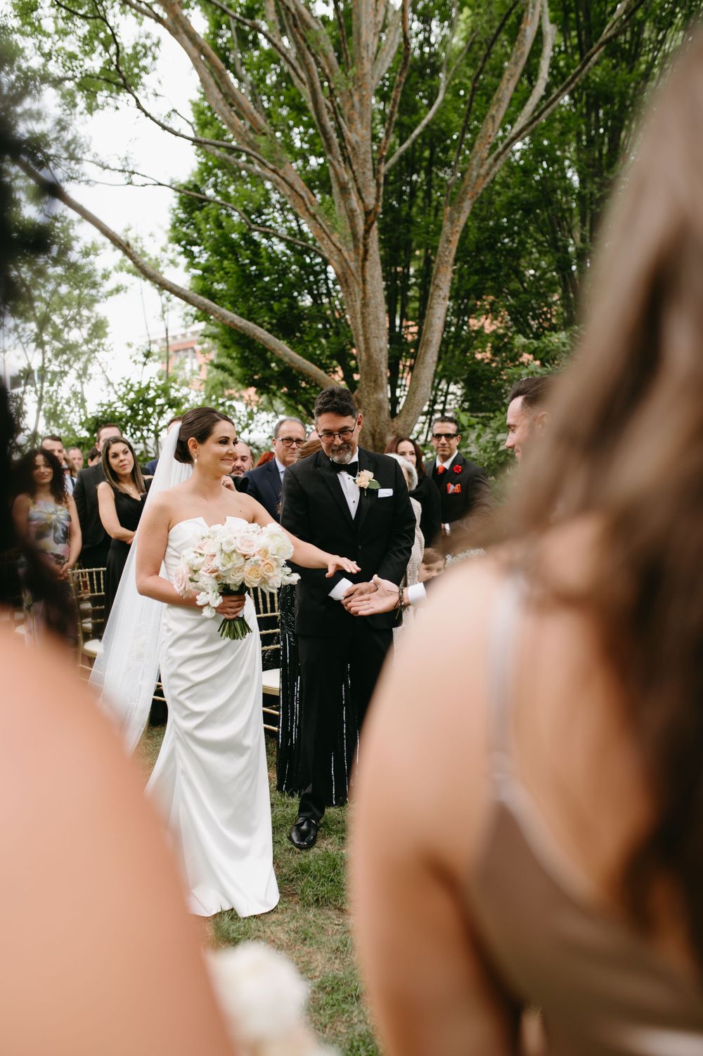 Wedding ceremony procession outdoors with guests watching from wooden chairs.